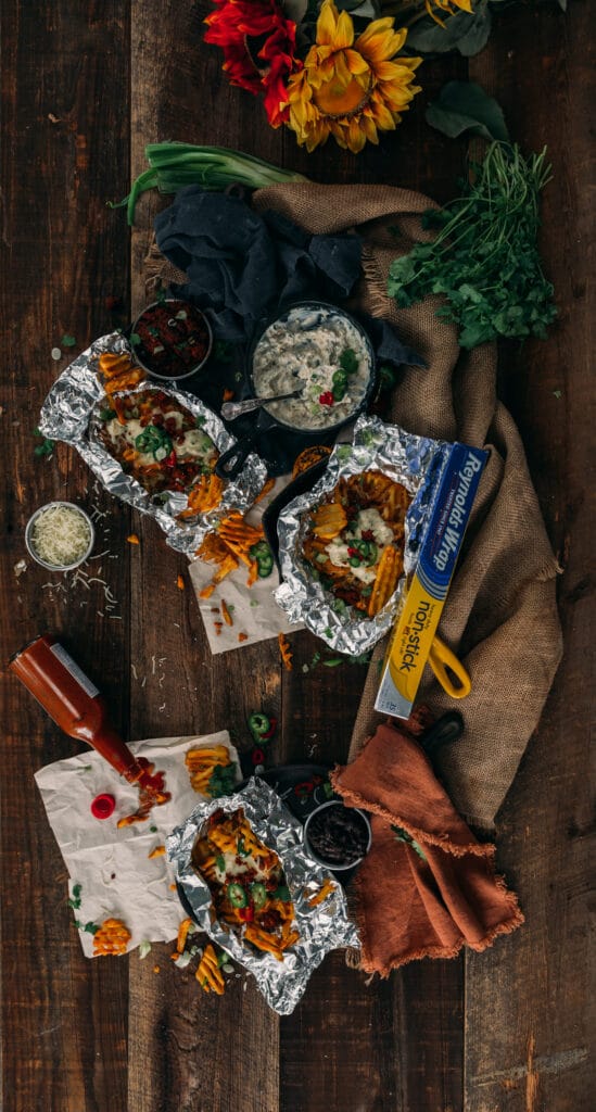 foto acima de batatas fritas waffle bagunçadas em pacotes de papel alumínio cobertos com molho cremoso de jalapeño com vários outros ingredientes espalhados 