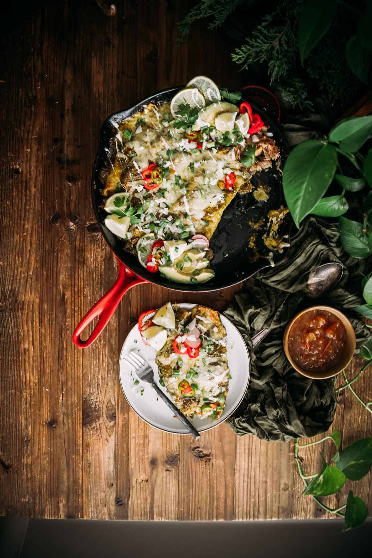 Uma frigideira de enchiladas cobertas com creme de leite, limão e pimenta em uma mesa de madeira, acompanhada por uma tigela de molho e pratos prontos para servir.