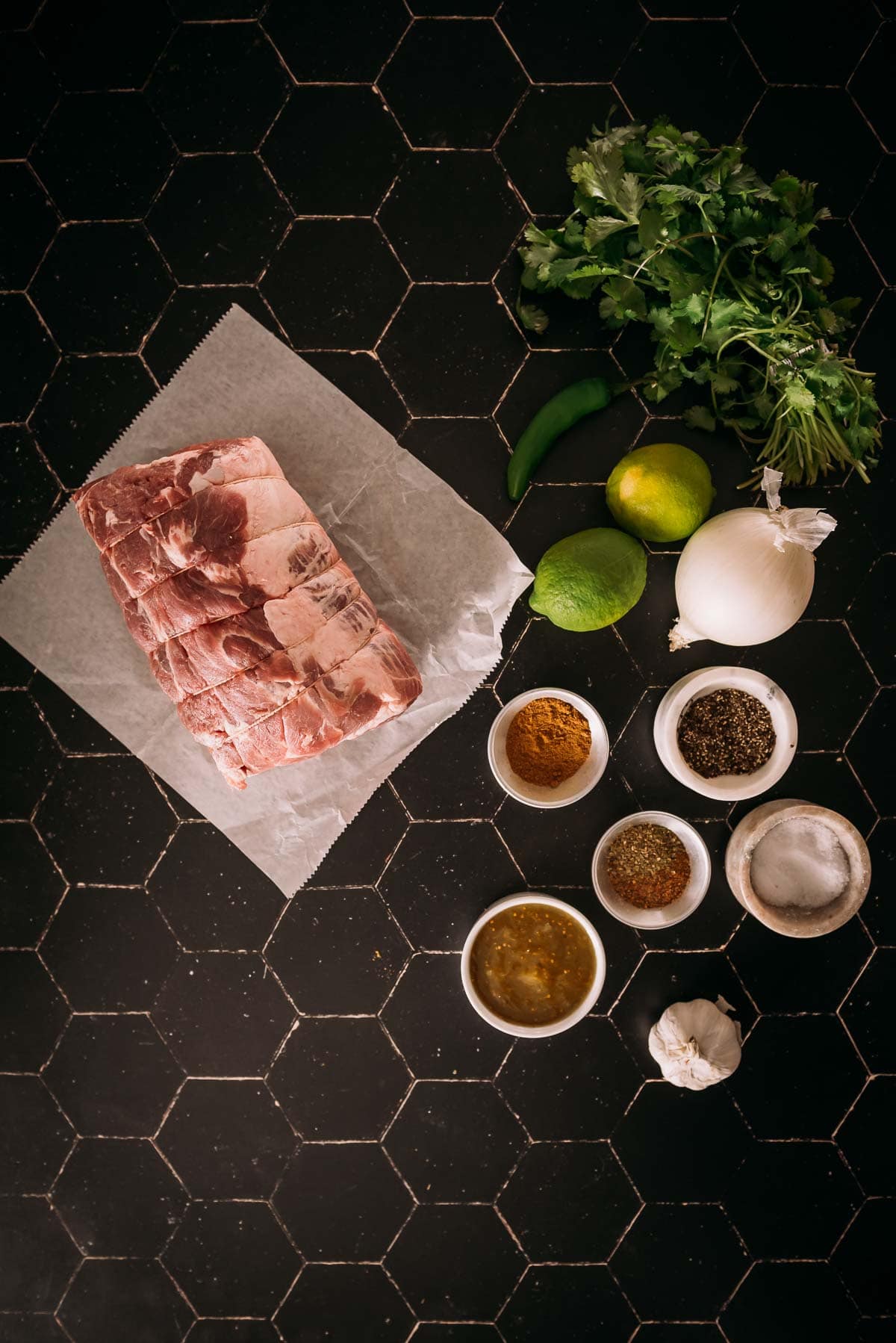 Ingredientes para uma receita dispostos em uma superfície de azulejos escuros, incluindo uma fatia de paleta de porco crua, ervas, especiarias, limão e cebola.