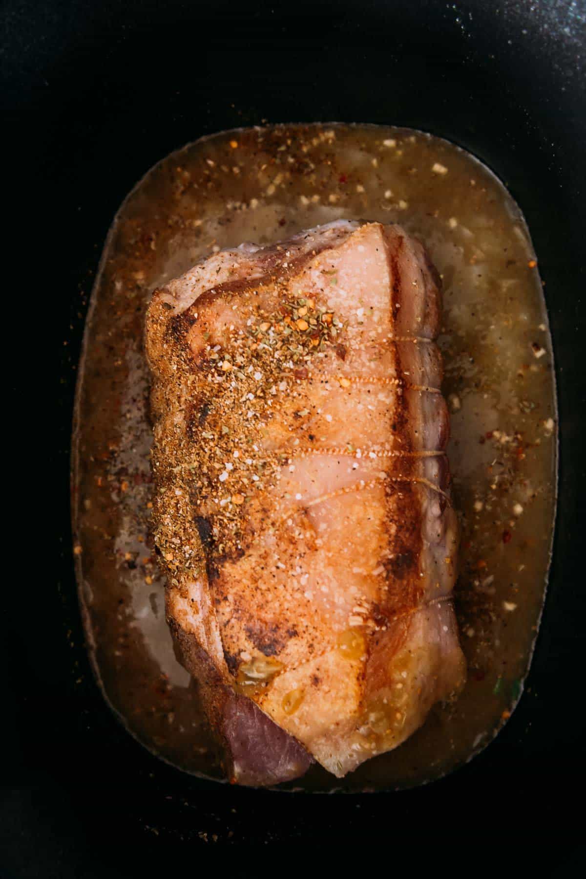 Um assado de porco temperado em panela elétrica com molho de enchilada verde com ervas e especiarias visíveis.