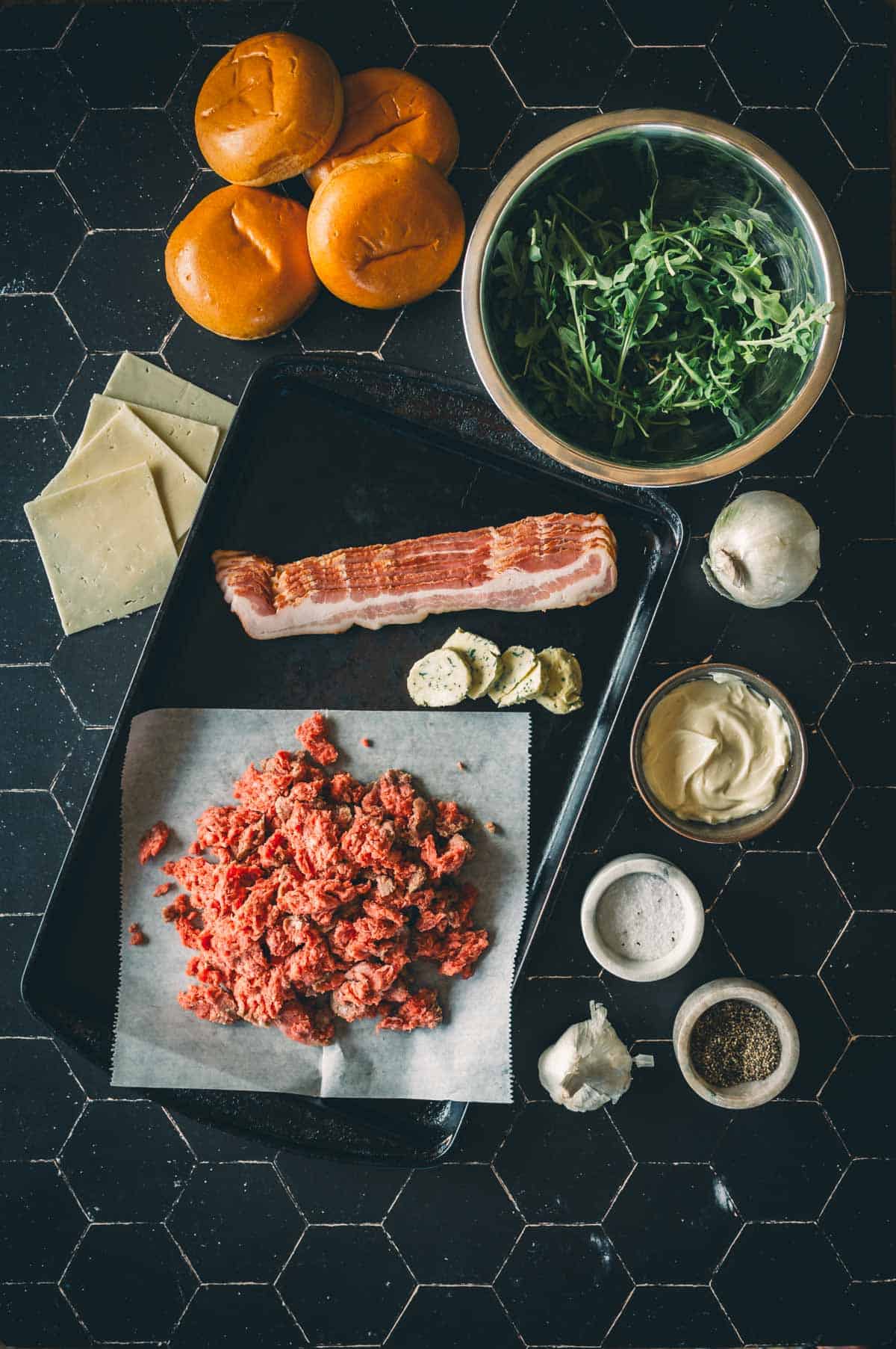 Ingredientes para fazer hambúrgueres, incluindo carne moída, bacon, fatias de queijo, pães de brioche, rúcula, alho, sal, pimenta, manteiga e maionese, dispostos sobre uma superfície preta.