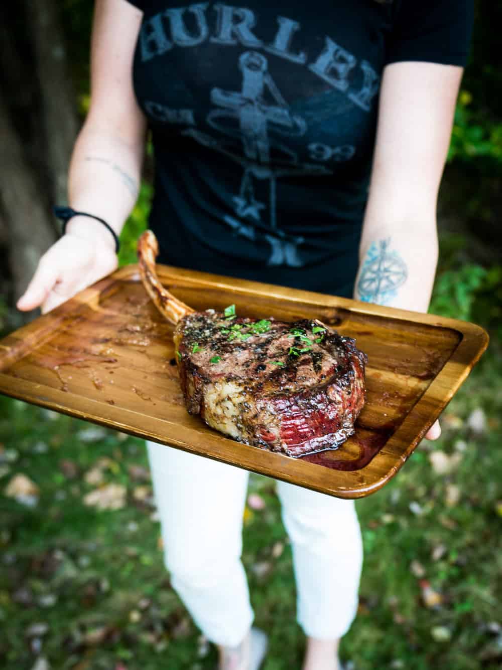 Uma pessoa vestindo camiseta preta e calça branca segura uma bandeja de madeira com um bife tomahawk grelhado e guarnecido com ervas.