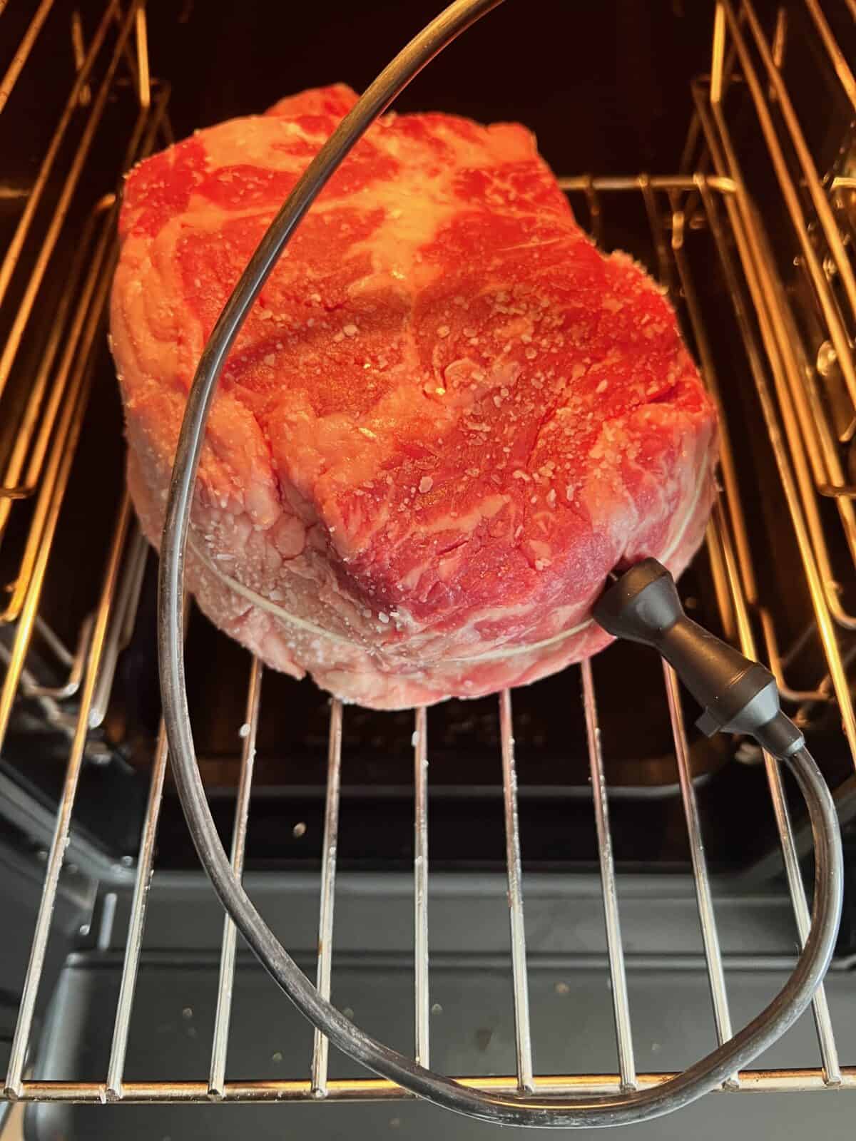 Um bife de cowboy cru com sal é colocado em um defumador em uma prateleira de metal, com uma sonda de termômetro de carne inserida nele.