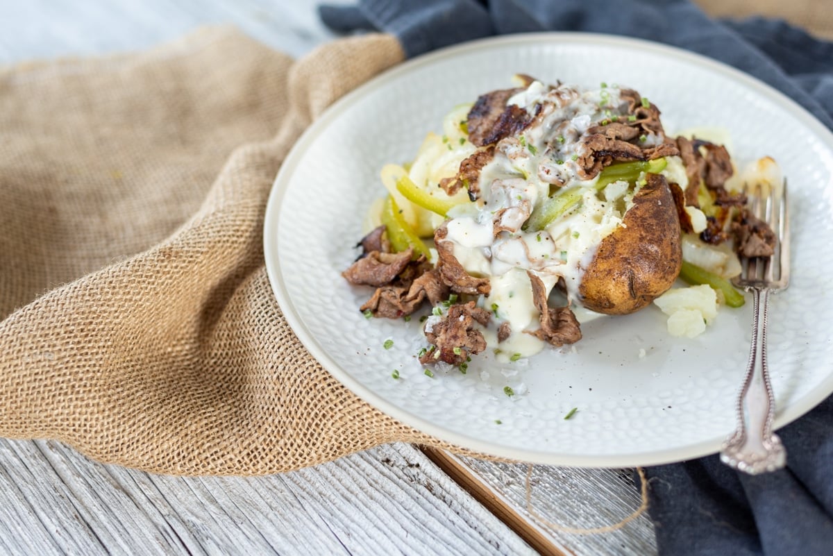 Uma batata assada carregada em um prato branco transbordando com molho cremoso de queijo, pimentão, cebola e bife cozido em fatias finas
