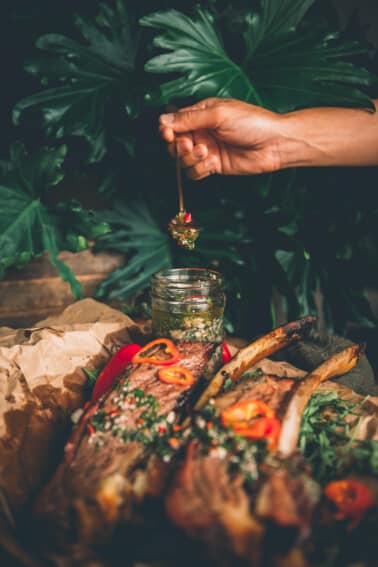 Mão segurando uma colher cheia de chimichurri sobre uma jarra com costelas de boi cobertas com molho em primeiro plano.