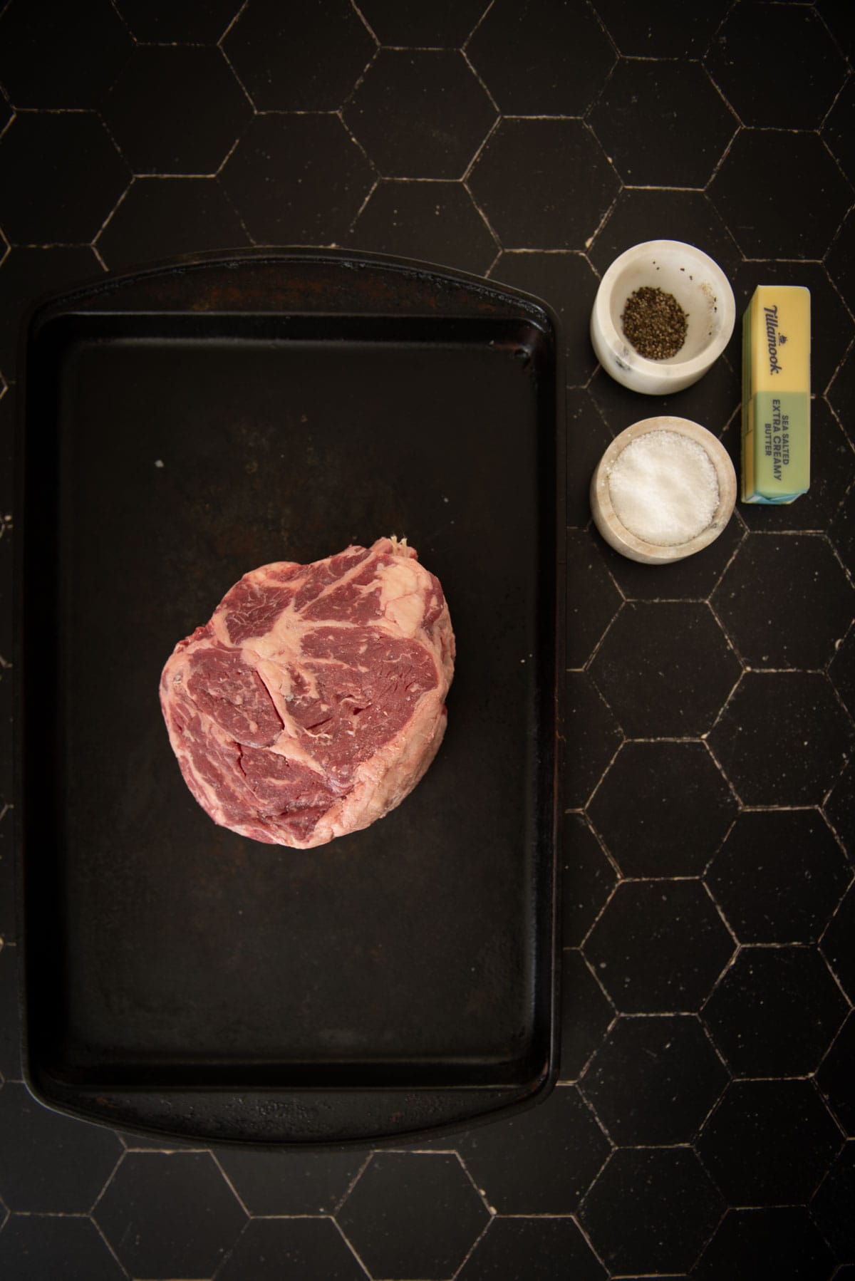 Um bife cru em uma assadeira preta com uma xícara de pimenta, uma xícara de sal e um pedaço de manteiga sobre uma superfície de azulejos hexagonais.