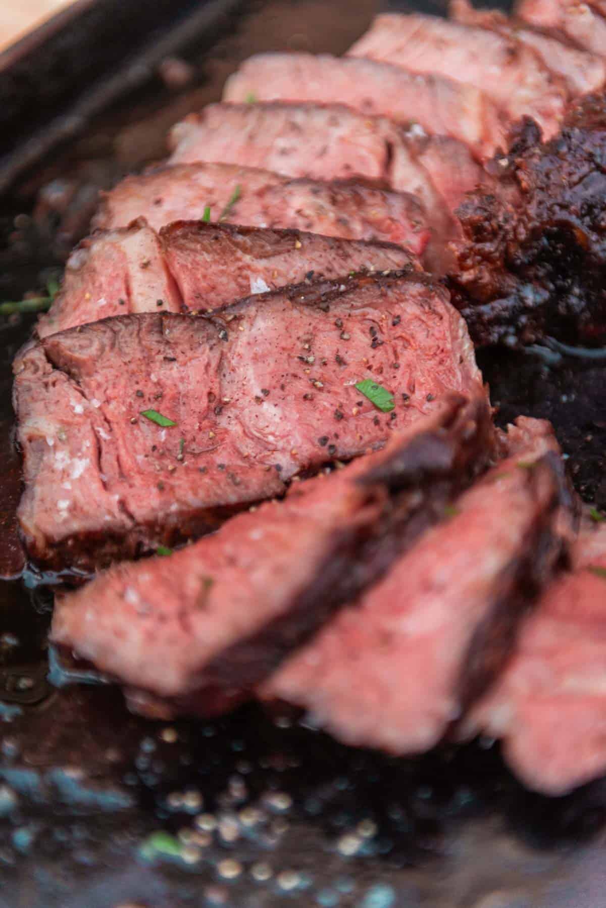 Close-up de lombo de cowboy fatiado e mal passado, temperado com ervas e pimenta em um prato preto.