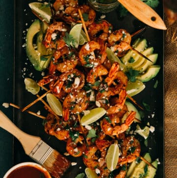 travessa de espetos de camarão grelhados servidos com molho barbecue, sobre rodelas de abacate, guarnecidos com rodelas de limão, cotija e chimichurri