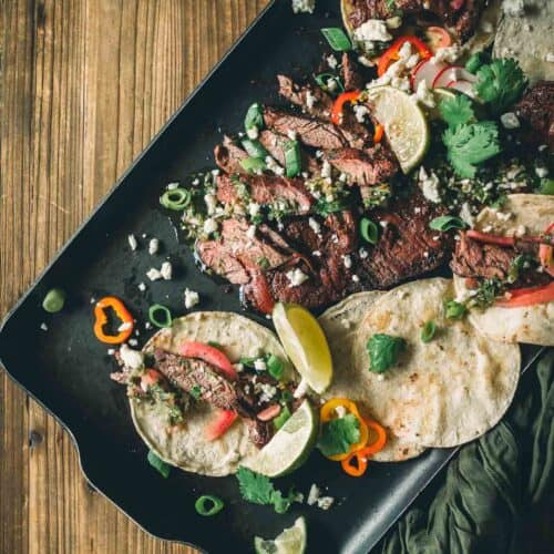 Uma bandeja com tacos de fraldinha grelhados, guarnecidos com rodelas de limão, coentro, cebolinha, queijo esfarelado e pimentão vermelho fatiado em tortilhas de farinha.