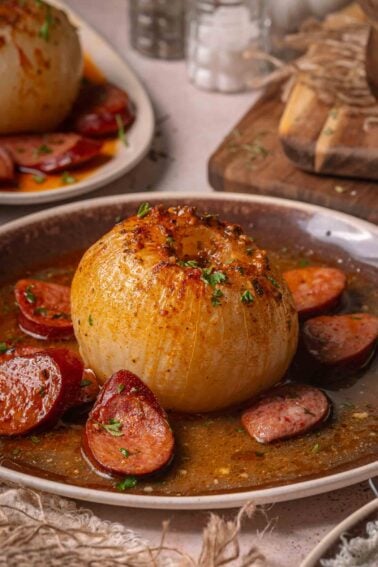 Uma cebola inteira servida em molho amanteigado com andouille fatiado em um prato marrom, guarnecido com ervas picadas.