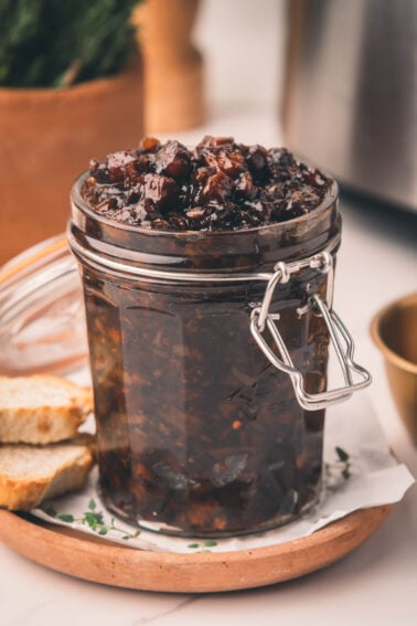 Uma jarra de vidro cheia de geleia de bacon fica em um prato com duas fatias de pão ao lado.