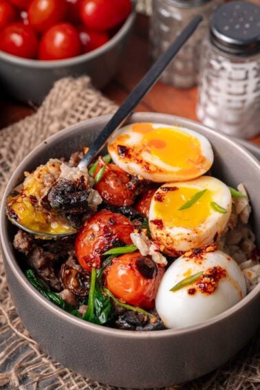 Uma tigela de tomates assados, verduras salteadas, carne moída e ovos cozidos cortados ao meio, guarnecidos com cebolinha. Uma colher está dentro da tigela.