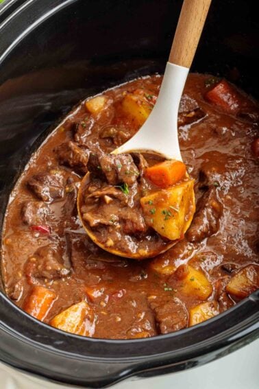 Uma concha de madeira tira ensopado de coração de boi com batatas e cenouras de uma panela elétrica, mostrando pedaços de carne e vegetais em um caldo marrom espesso.