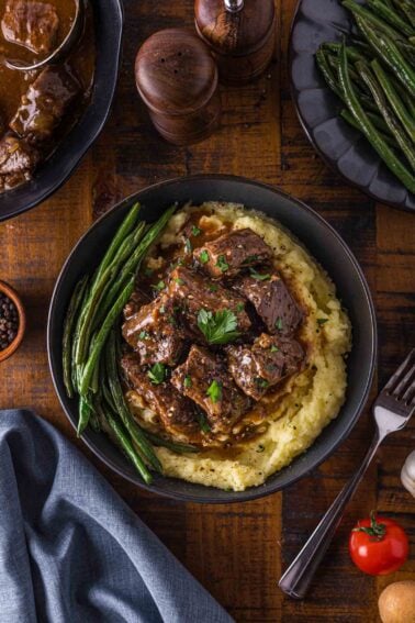 Uma tigela de purê de batata coberto com ponta de carne ao molho, guarnecido com salsa e servido com feijão verde; lados e utensílios adicionais estão sobre uma mesa de madeira.