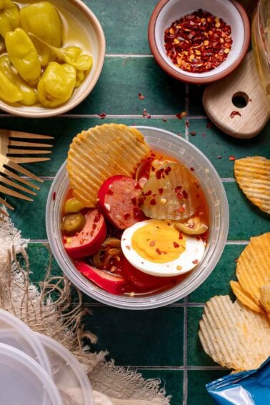 A tigela picante viral com picles fatiados, ovos cozidos, linguiça vermelha, pimentão banana e batata frita, coberta com flocos de pimenta vermelha em uma superfície de ladrilho verde.