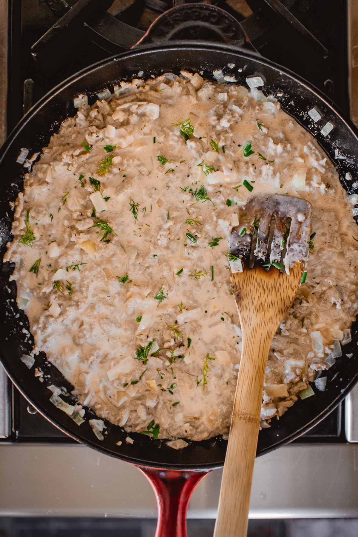 Uma frigideira com peru cremoso moído e molho de ervas, com cebola picada e ervas, com uma espátula de madeira apoiada na borda.