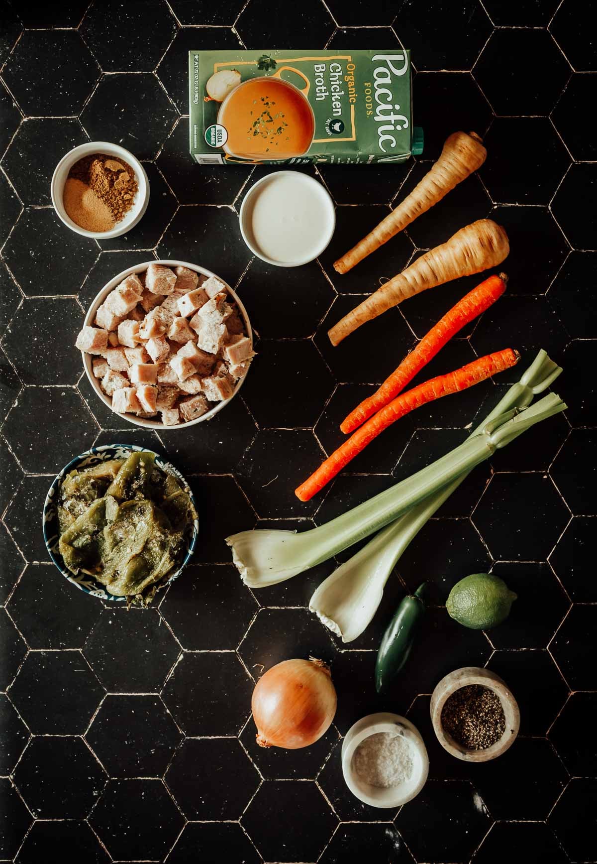 Os ingredientes em uma superfície de ladrilho preto incluem caldo de galinha, sobras de peru, nabo, cenoura, aipo, pão em cubos, pimenta verde, cebola, jalapeño, limão e temperos em tigelas pequenas.