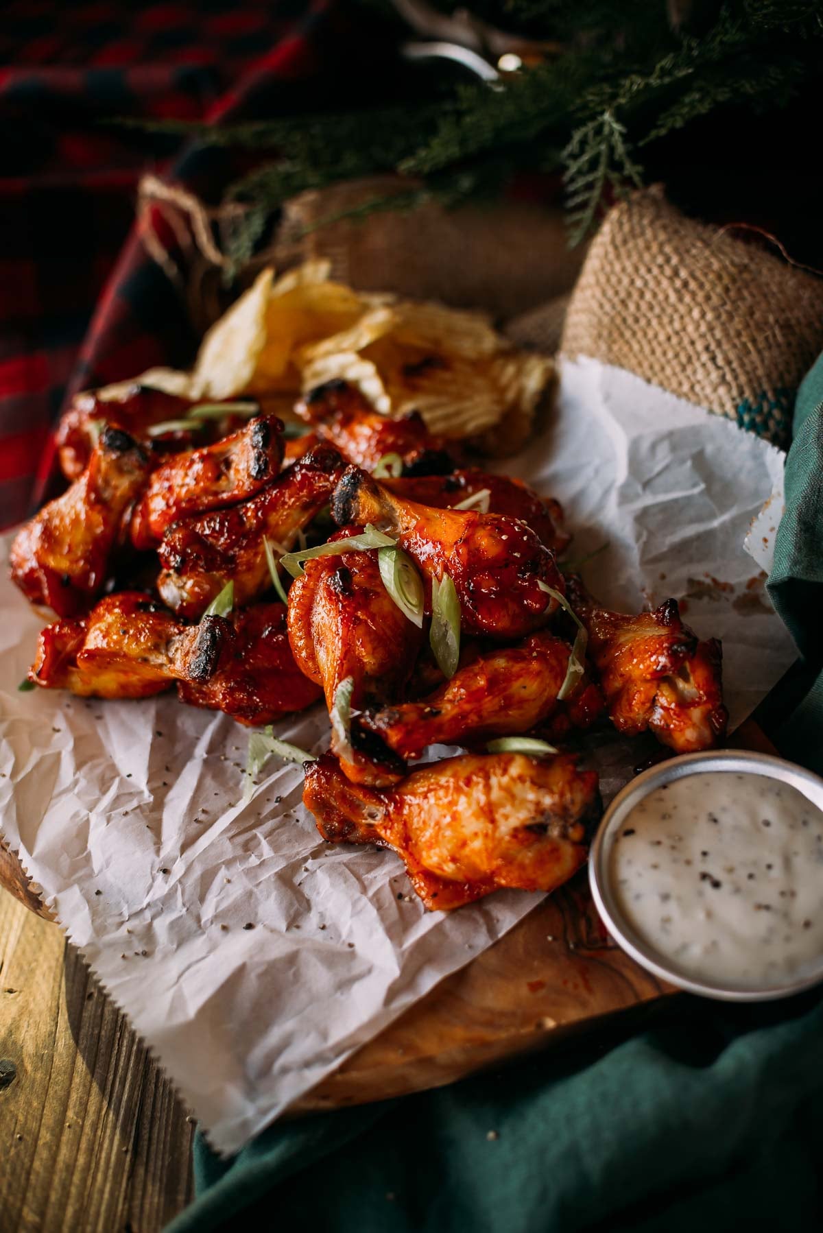 Uma porção de asas de sriracha com mel picante guarnecidas com cebolinha, colocadas em papel manteiga amassado, com molho e batatas fritas ao fundo.