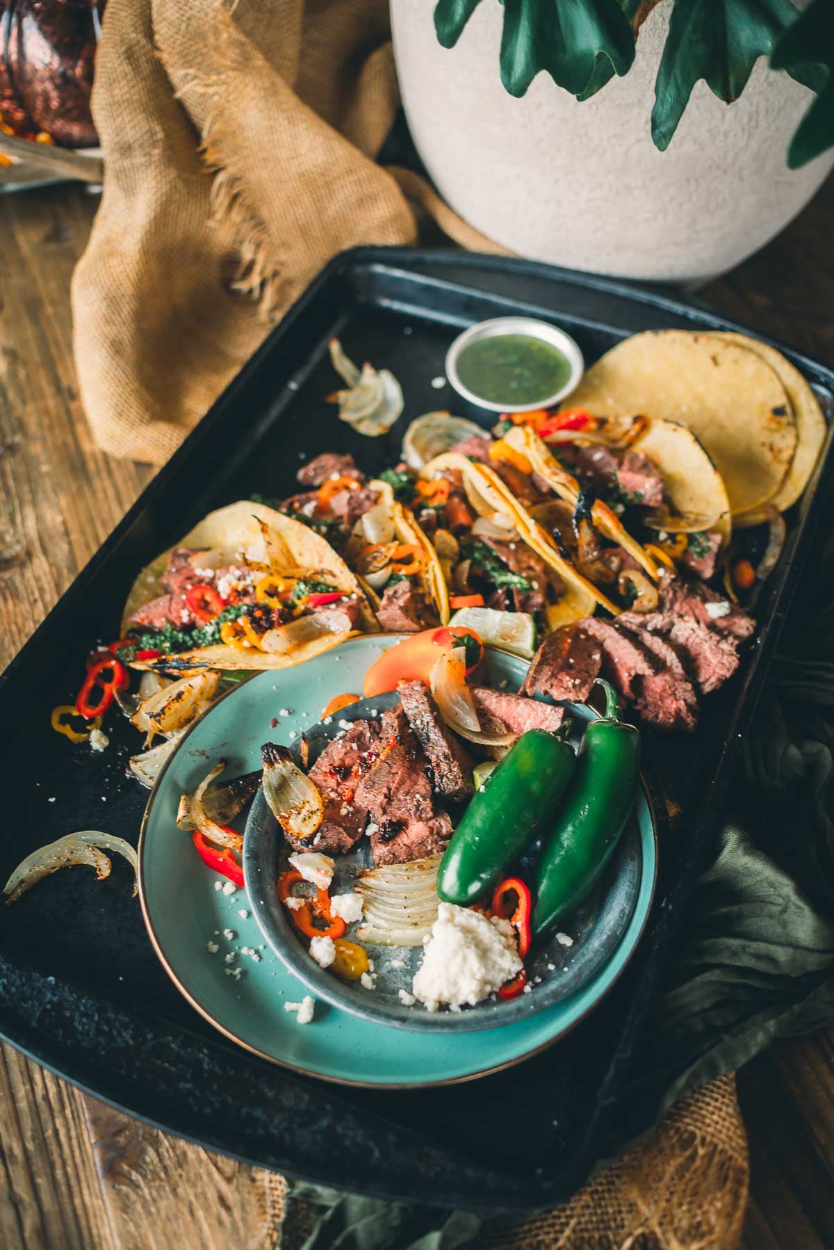 Um prato com tacos de bife fatiados, jalapeños e queijo, cercados por coberturas de taco em uma bandeja. Decorado com cebola e pimentão, com uma tigela pequena de chimichurri.