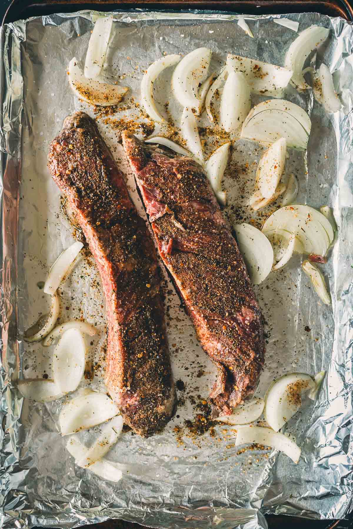 Dois bifes de carne temperada e cebolas fatiadas em uma assadeira forrada com papel alumínio.