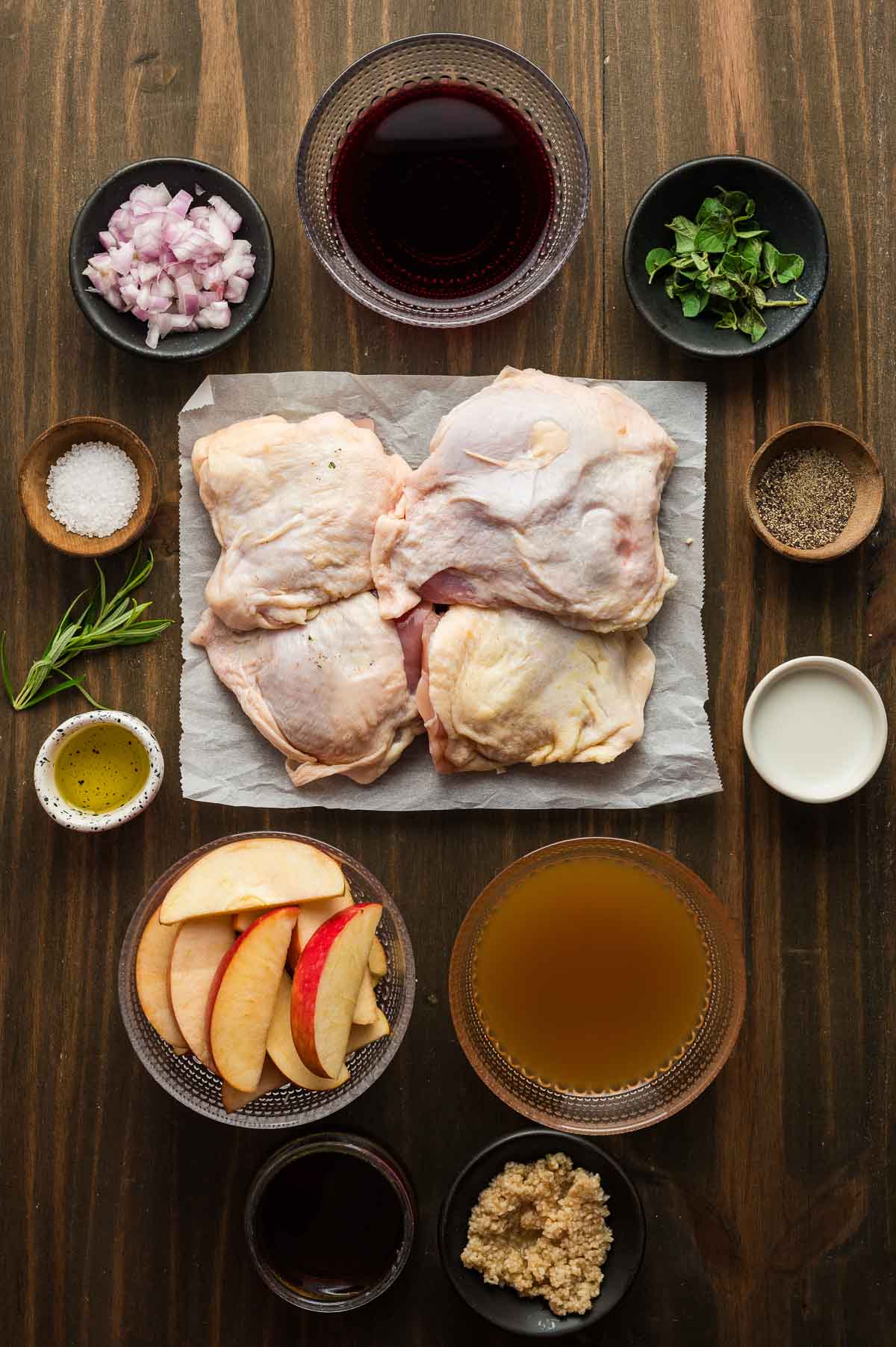 Coxas de frango cru em pergaminho rodeadas por fatias de maçã, cebolinha picada, alho picado, ervas, vinho tinto, cidra de maçã, azeite, sal e pimenta sobre uma superfície de madeira.