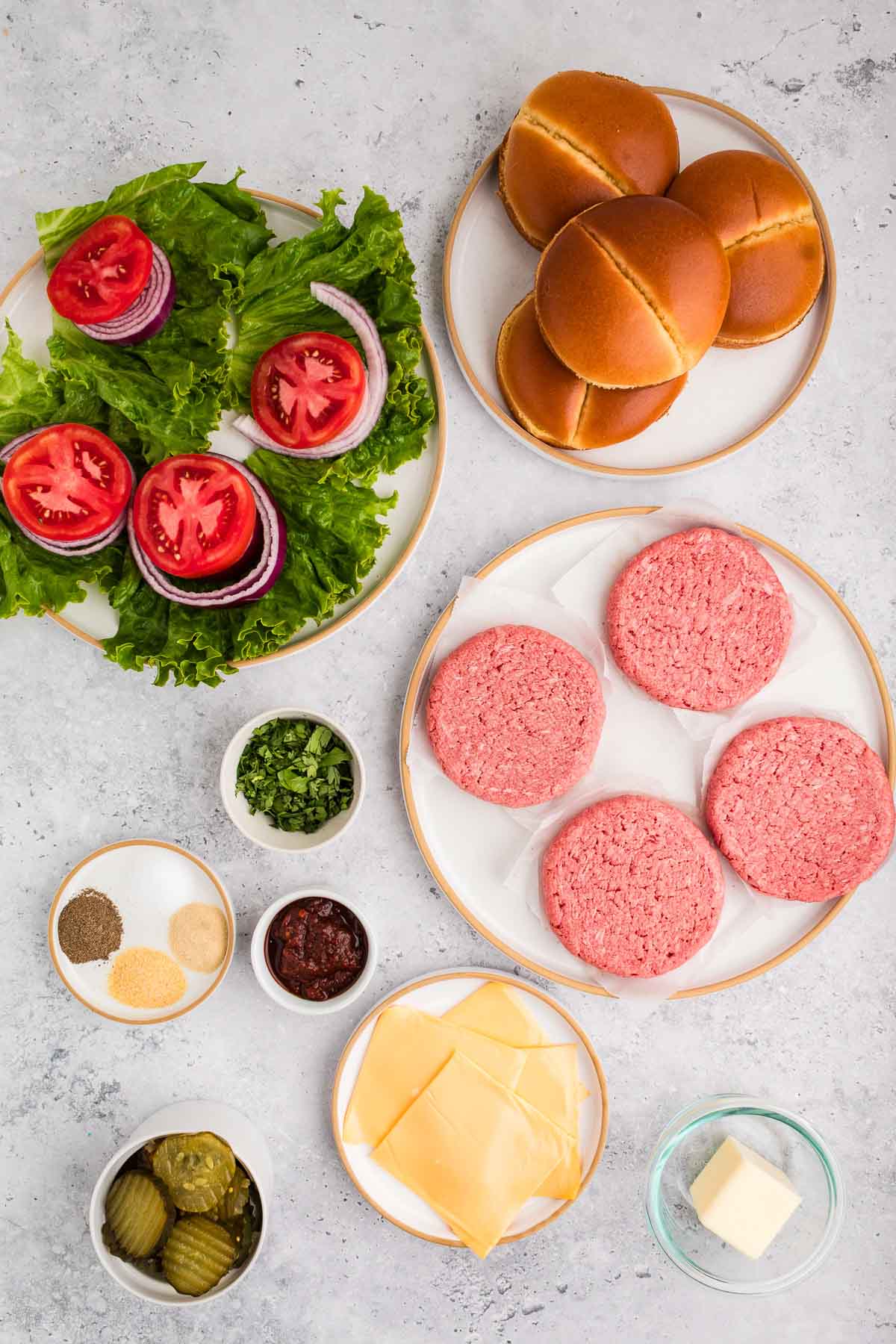 Ingredientes do hambúrguer dispostos sobre uma mesa, incluindo hambúrgueres de carne crua, pães, alface, tomate, cebola, queijo fatiado, picles, temperos, ervas picadas, molho e uma tigela pequena de manteiga.