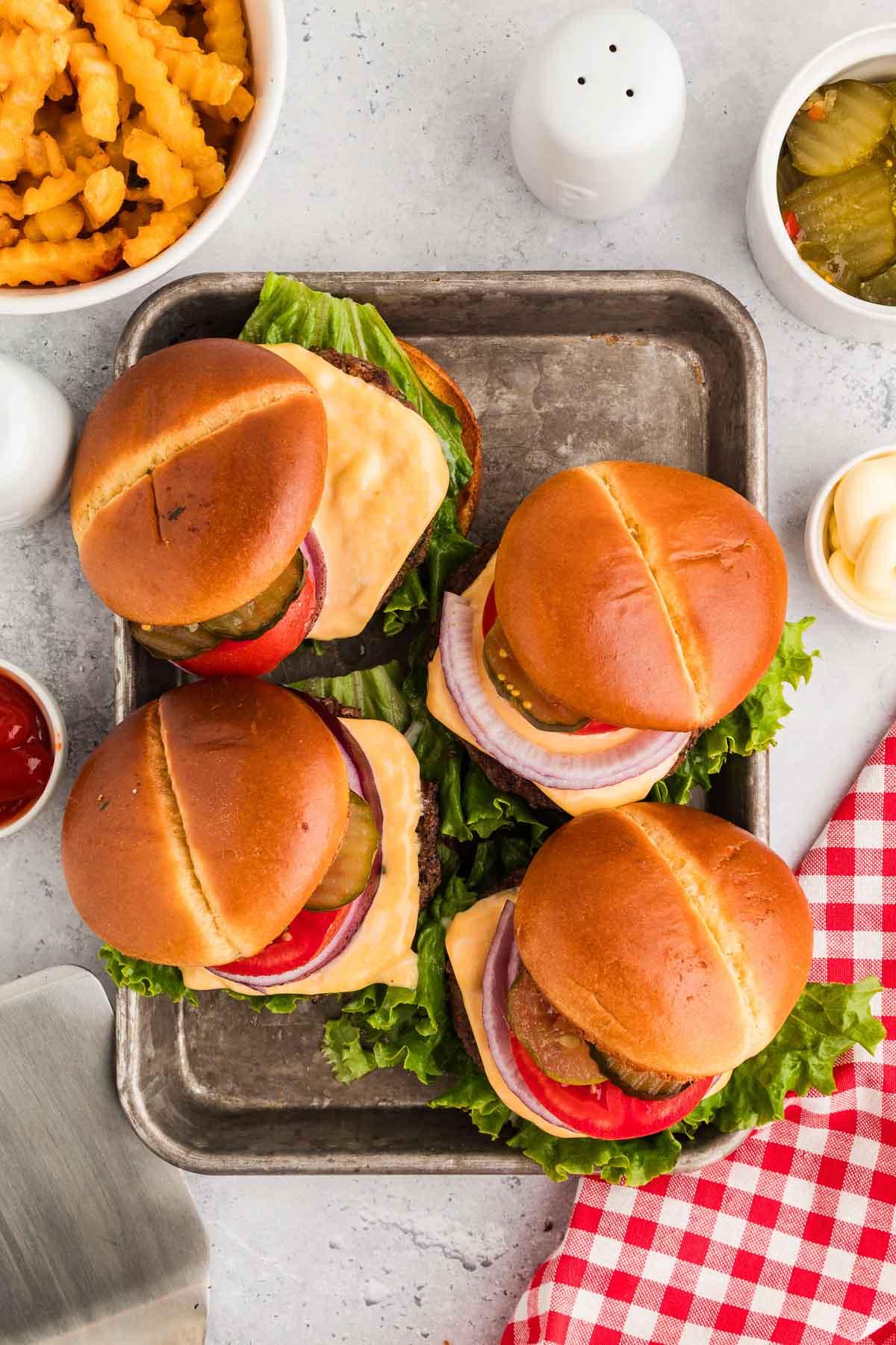 Quatro hambúrgueres de manteiga cowboy com alface, tomate, picles, cebola e queijo em uma bandeja, rodeados de batatas fritas amassadas, picles, ketchup, maionese e um guardanapo xadrez vermelho.