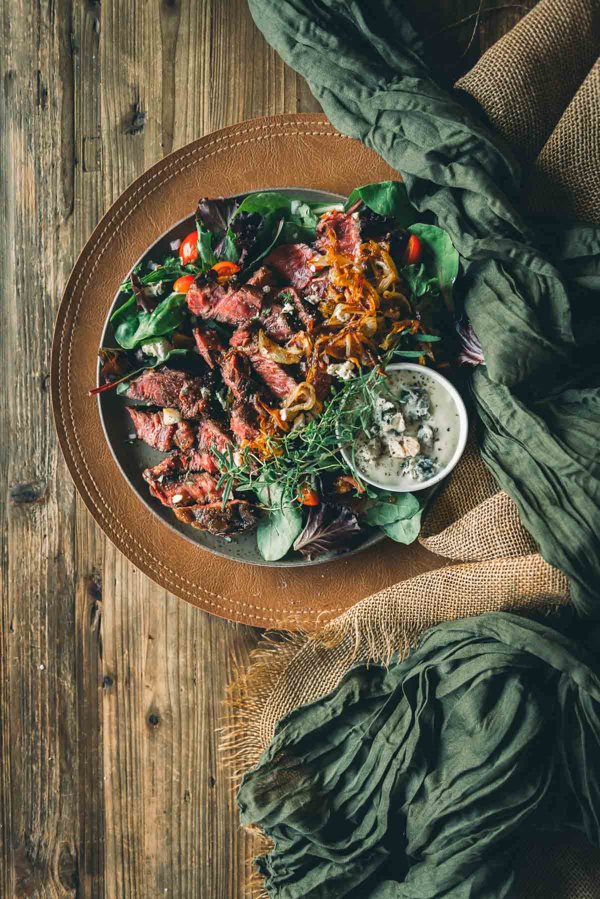 Um prato de bife fatiado com verduras mistas com cebola assada, tomate cereja, microgreens e uma tigela pequena de molho cremoso, colocado sobre uma mesa de madeira rústica.