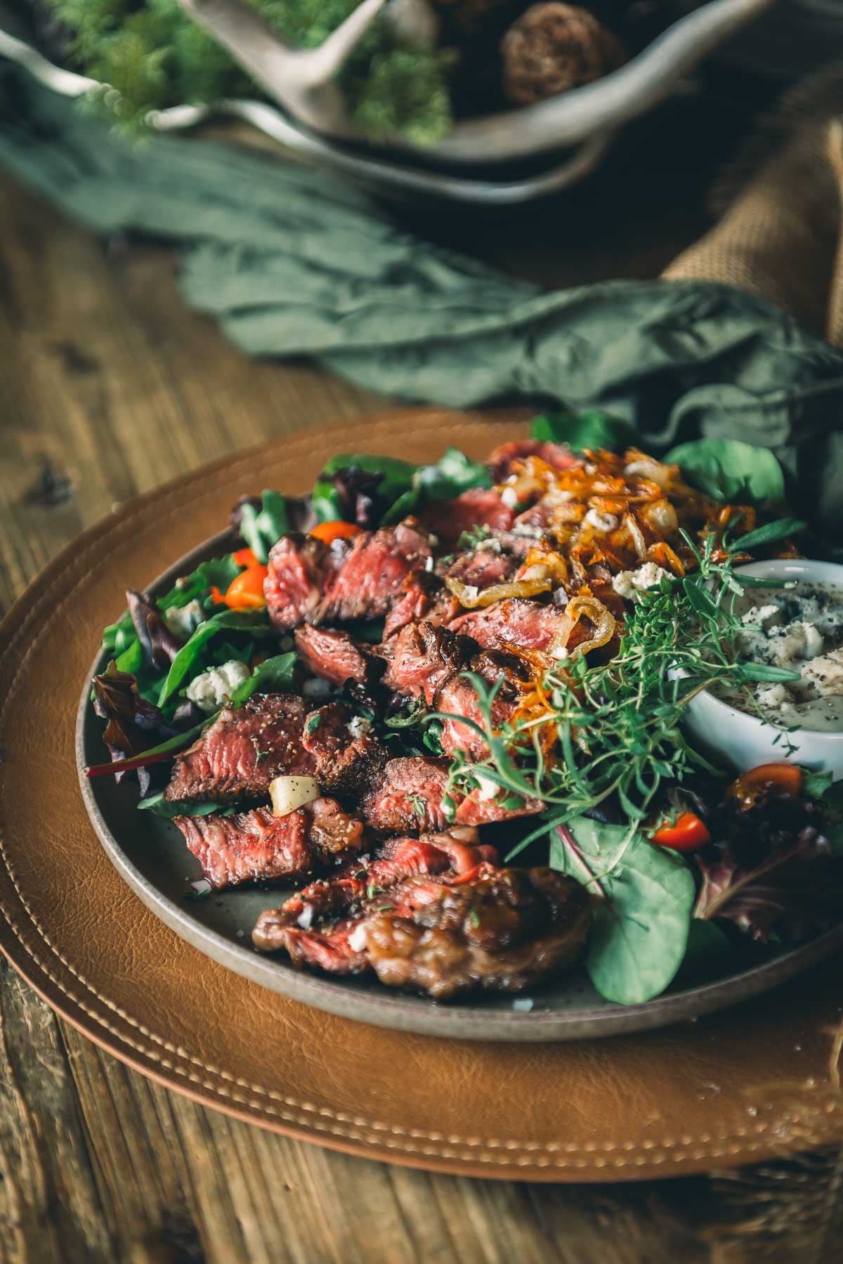 Um prato de bife grelhado fatiado servido em uma mistura de verduras com cebola caramelizada, ervas frescas e um pequeno prato de molho de queijo azul em uma mesa de madeira.