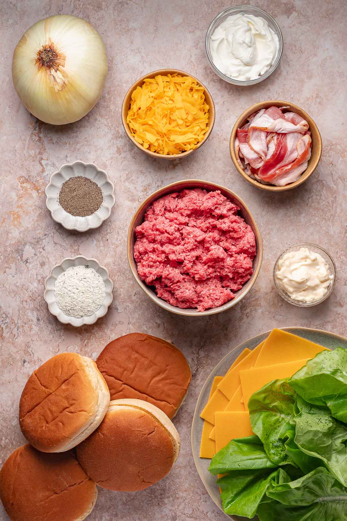 Vista de cima para baixo dos ingredientes do hambúrguer, incluindo carne moída, pães, alface, queijo cheddar, bacon, queijo ralado, cebola, pimenta preta, sal, maionese e creme de leite em uma bancada.