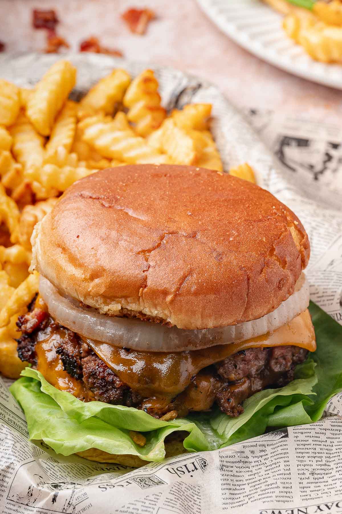 Cheeseburger com alface, cebola e queijo derretido no pão, servido com batatas fritas amassadas em papel tipo jornal.