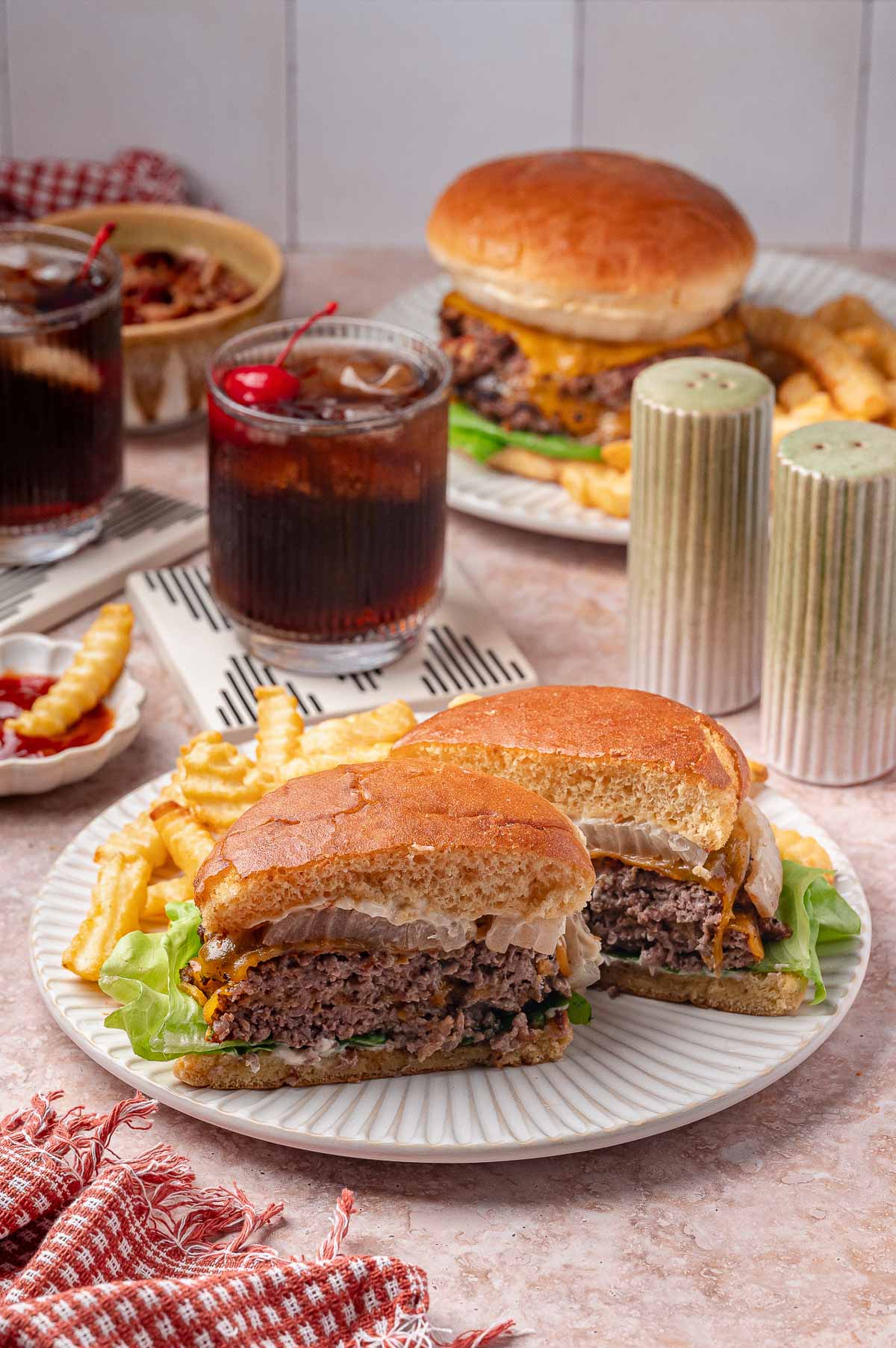 Um prato com um cheeseburger cortado ao meio e batatas fritas amassadas está em primeiro plano, com dois refrigerantes, outro hambúrguer, mais batatas fritas e condimentos ao fundo.