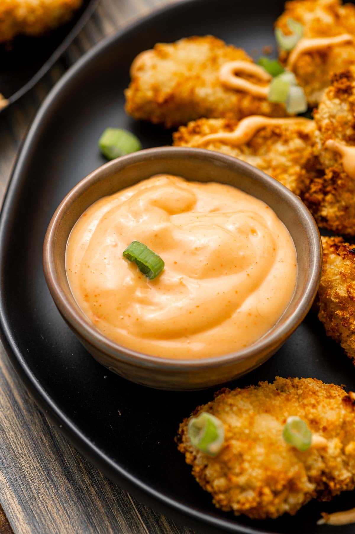 Uma pequena tigela de molho cremoso bang bang guarnecido com cebola verde picada, servido em um prato preto com aperitivos empanados e assados.