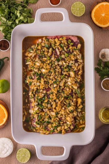 Uma assadeira branca contendo um pedaço de carne marinado em uma mistura de cebola picada, ervas e frutas cítricas, rodeado por ingredientes frescos em uma superfície marrom.