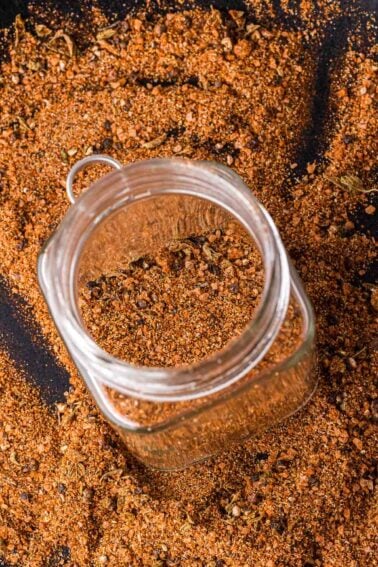 Uma jarra de vidro cheia de uma mistura de temperos de carne assada é cercada por mais do mesmo tempero espalhado em uma superfície escura.