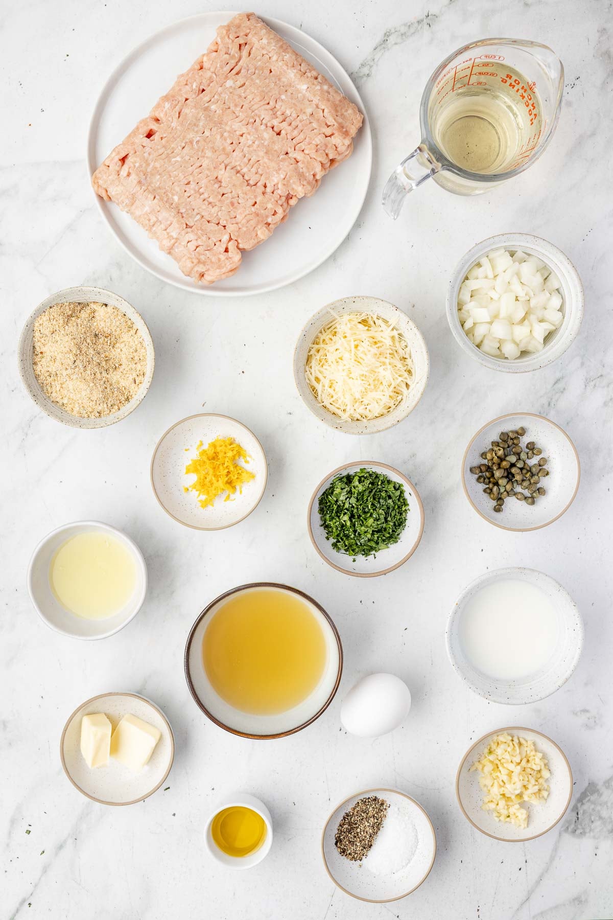 Ingredientes para Almôndegas de Frango Piccata expostos em superfície branca, incluindo carne moída, cebola picada, pão ralado, queijo ralado, ervas, alcaparras, raspas de limão, ovo, caldo, manteiga, óleo, leite e temperos.