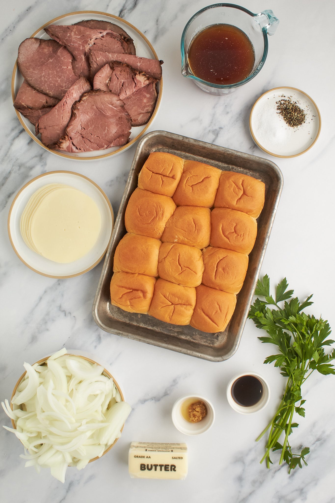 Ingredientes para sliders franceses dispostos em uma superfície de mármore, incluindo pãezinhos, rosbife, queijo provolone, cebola, manteiga, caldo, molho inglês, temperos e salsa fresca.