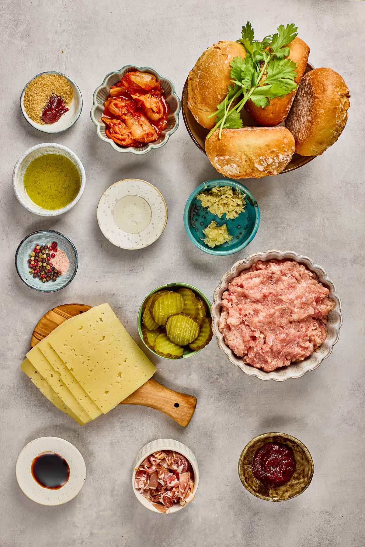 Uma visão aérea dos ingredientes do hambúrguer, incluindo carne moída, pãezinhos, fatias de queijo, picles, molhos, temperos, ervas e pequenas tigelas com outros condimentos em uma superfície leve.