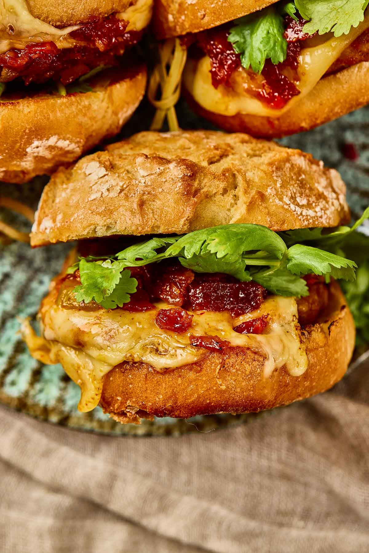 Close de um hambúrguer de frango Gochujang com queijo derretido e geléia de bacon kimchi em um pão rústico, colocado em um prato de cerâmica.