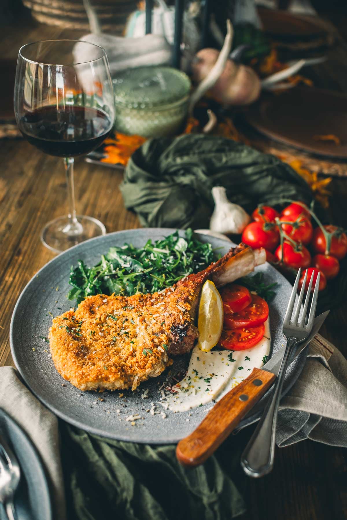 Uma refeição preparada com vitela à milanesa empanada com rodela de limão, tomate fatiado, folhas verdes e uma taça de vinho tinto em uma mesa de madeira.