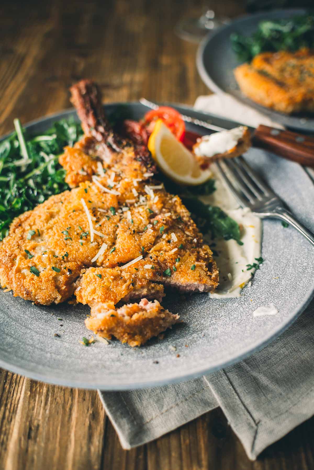 Um prato de costeleta de vitela à milanesa crocante servida com verduras, tomate cereja, rodela de limão e guarnecida com queijo ralado em uma mesa de madeira. 