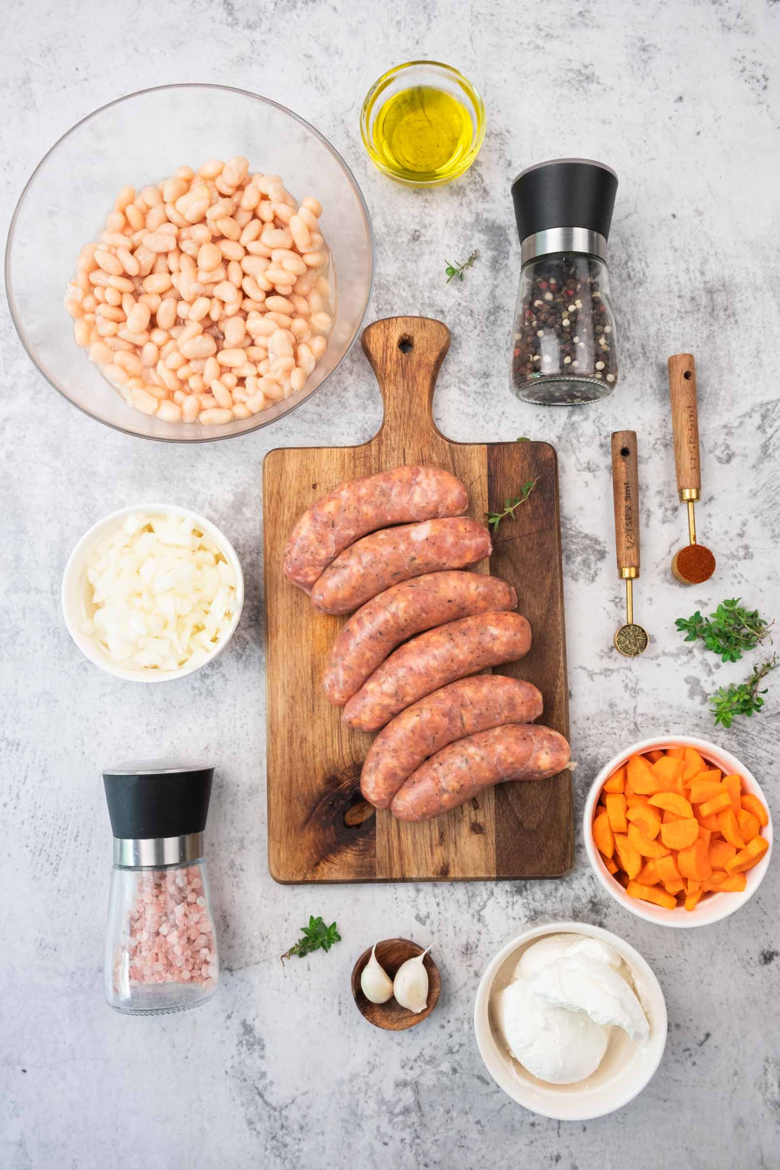 Vista de cima para baixo de salsichas cruas sobre uma tábua de corte, cercadas por tigelas de feijão branco, cenoura picada, cebola, alho, especiarias, ervas, azeite e moedores de sal e pimenta.