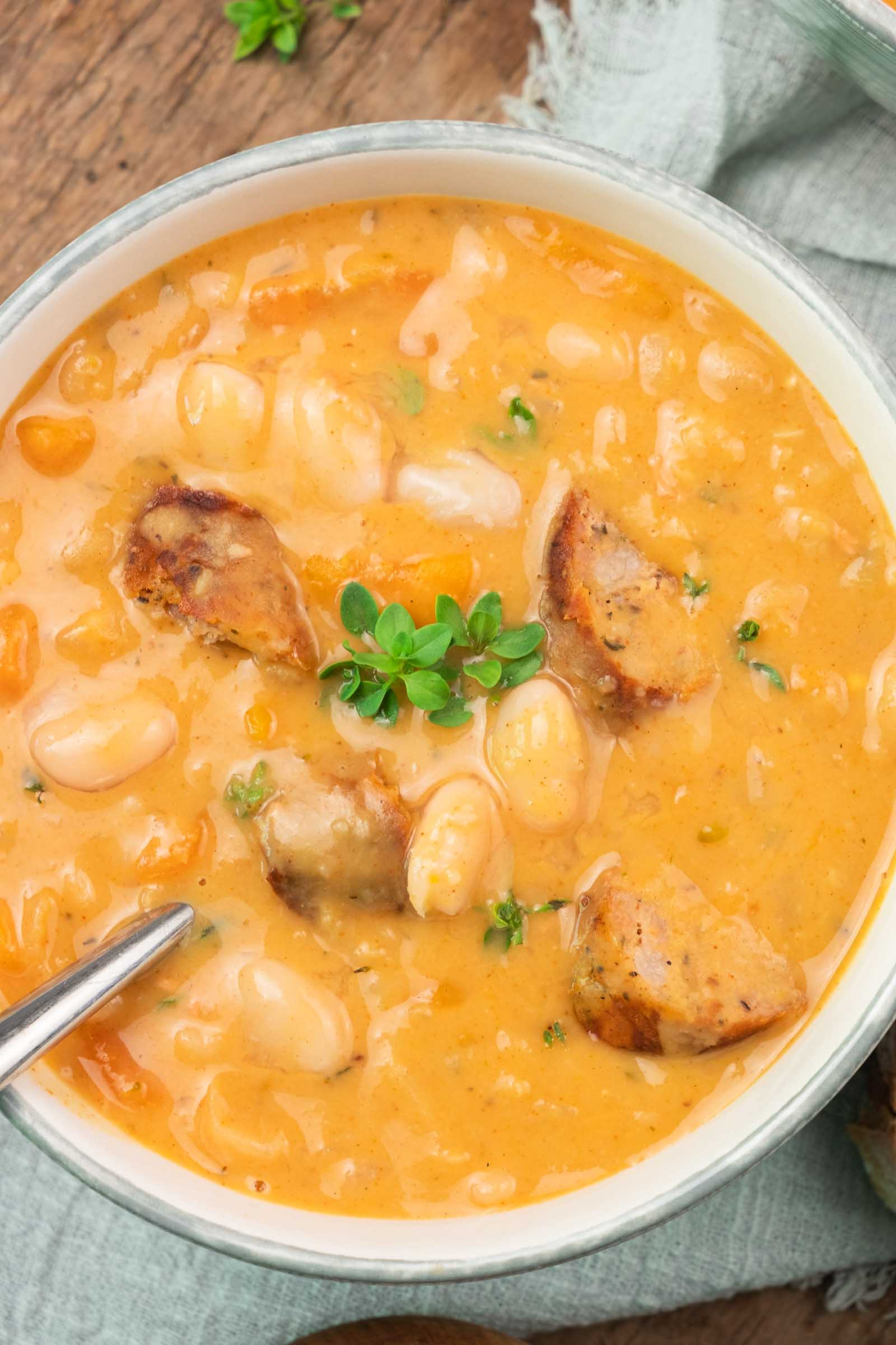 Uma tigela de sopa cremosa de feijão branco com fatias de salsicha, decorada com ervas frescas, repousa sobre uma superfície de madeira com uma colher.