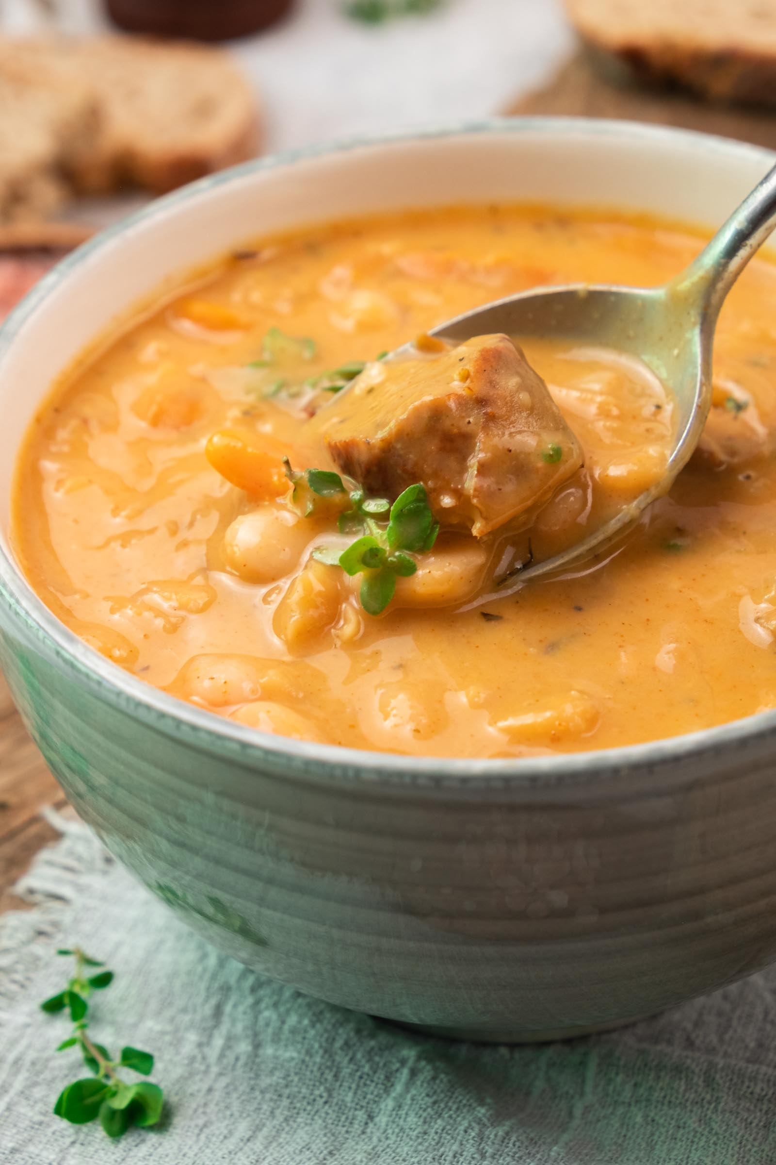 Close de uma tigela de sopa espessa de feijão branco com pedaços de linguiça, guarnecida com ervas frescas, e uma colher tirando um pedaço de carne da sopa.