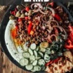 Um prato de cordeiro moído grego com fatias de pepino, cebola, tomate, azeitonas, pão pita e ricota batida, exposto sobre uma mesa de madeira. Leituras de texto "Como fazer cordeiro moído grego.