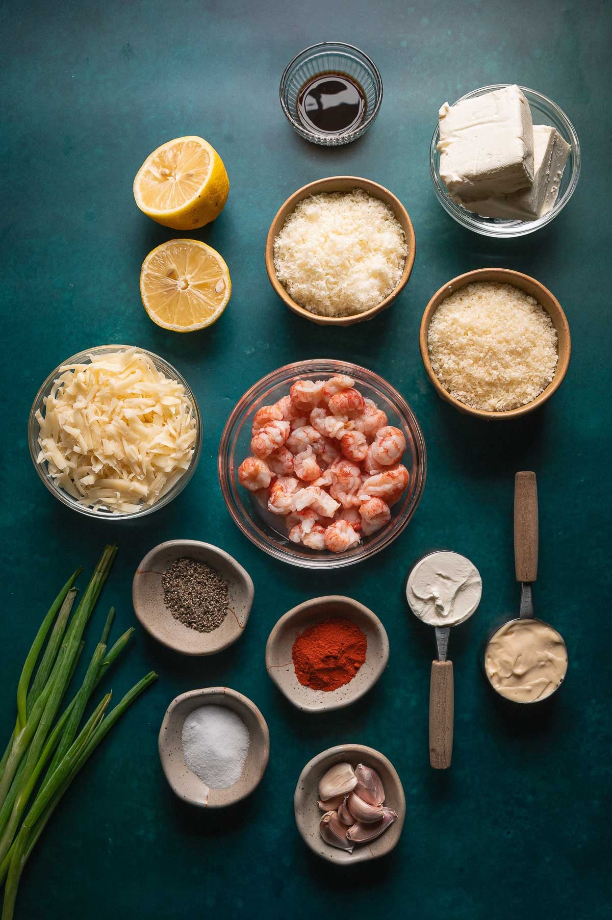 Vista de cima para baixo de vários ingredientes em tigelas e pratos, incluindo lagosta cozida, queijos, temperos, metades de limão, cebolinha e molhos cremosos em uma superfície verde escura.