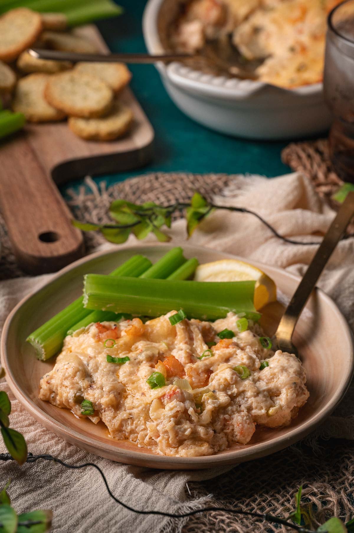 Um prato com molho cremoso de lagosta quente guarnecido com cebolinha, servido junto com talos de aipo e uma rodela de limão, com biscoitos e mais molho ao fundo.