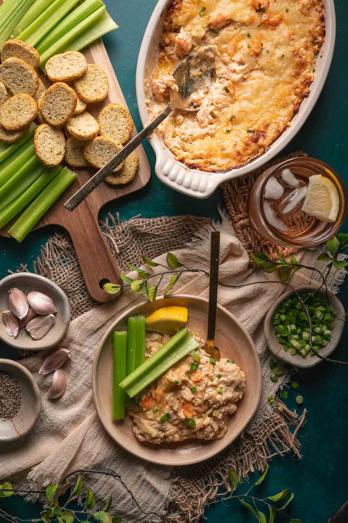 Uma tigela de molho cremoso de lagosta quente com talos de aipo e uma rodela de limão, ao lado de um prato de molho assado, fatias de pão torrado, um copo de bebida gelada, cebola verde fatiada, alho e pimenta.