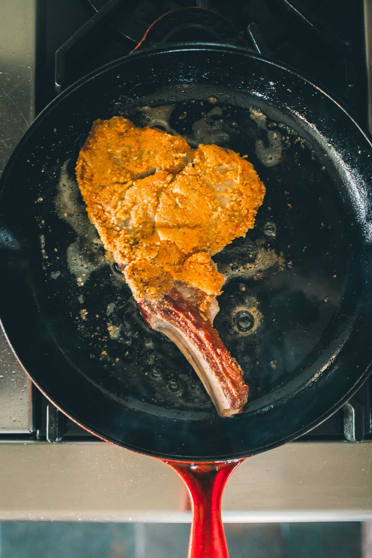 Uma costeleta de vitela temperada está sendo frita em uma frigideira preta de cabo vermelho no fogão.