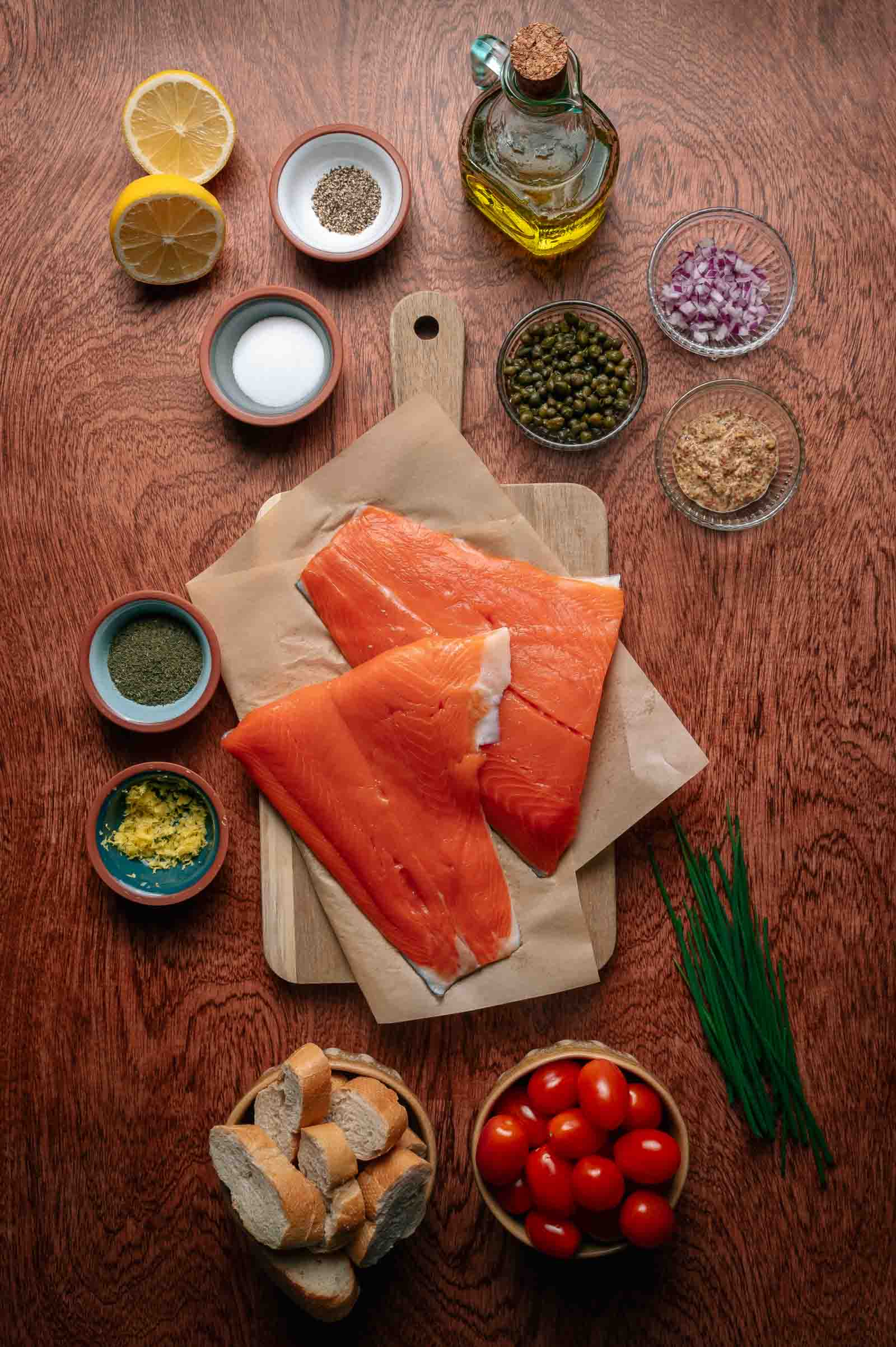 Dois filés de salmão cru em papel manteiga com tigelas de cebola picada, alcaparras, temperos, metades de limão, tomate cereja, fatias de pão, azeite e cebolinha fresca dispostas sobre uma mesa de madeira.